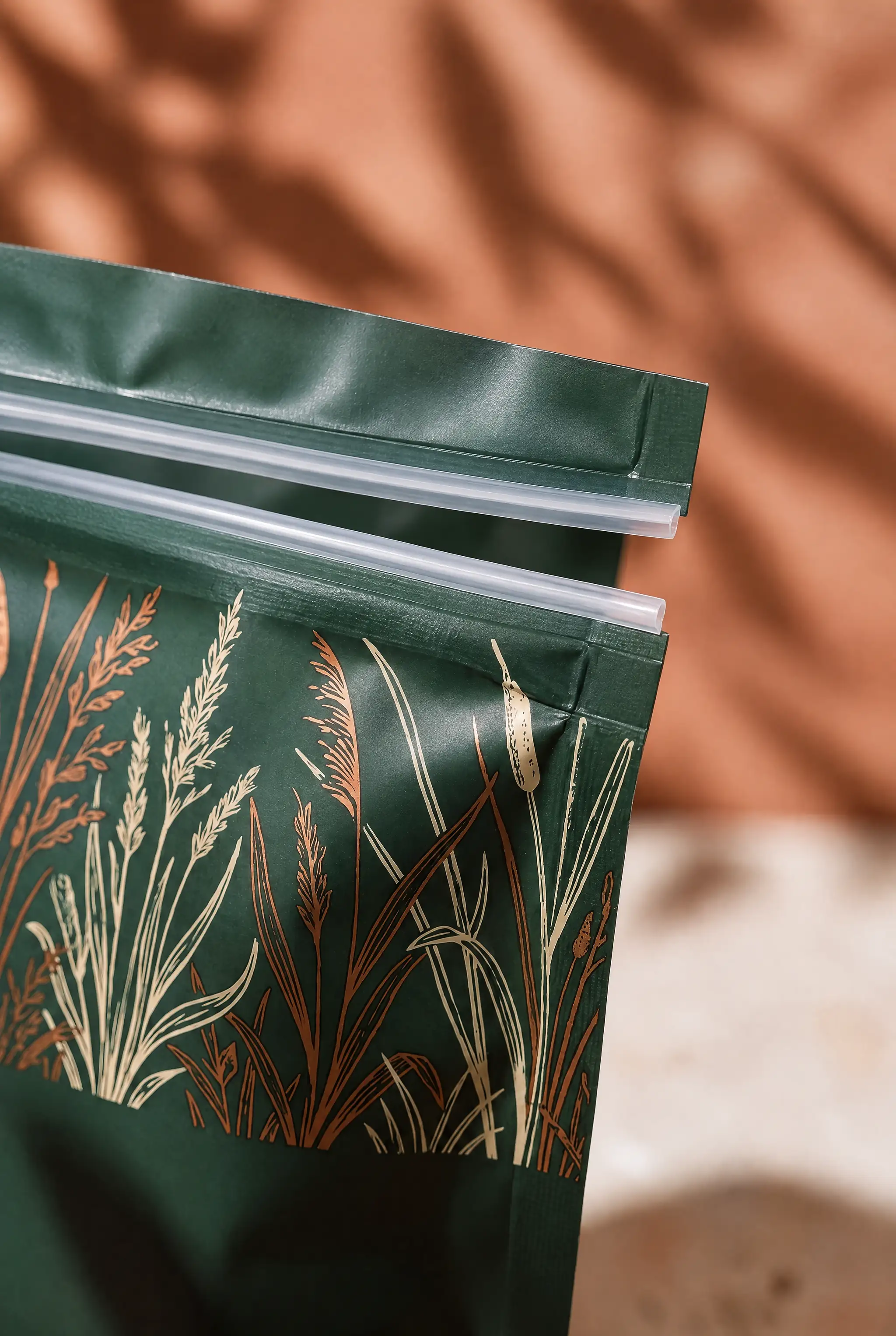 Macro view of the top of a stand-up pouch, where a wavy line cut through the film has parted into two scalloped edges with the background showing through the gap, above a panel printed with copper and cream grasses.