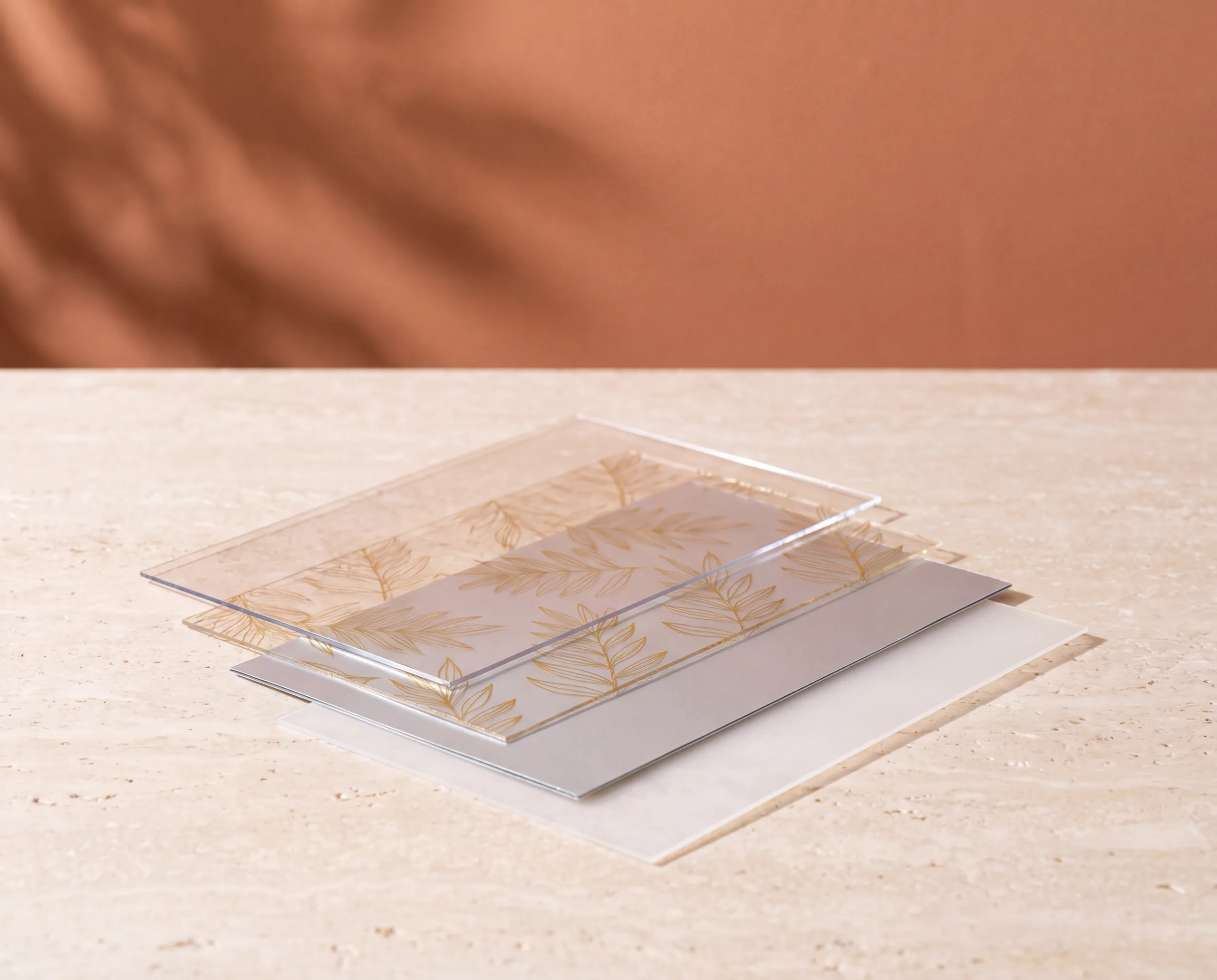 Four thin sheets of packaging film stacked on a stone table — a clear glossy outer sheet, a sheet printed in gold, a matte silver barrier sheet and a milky white sealing sheet.